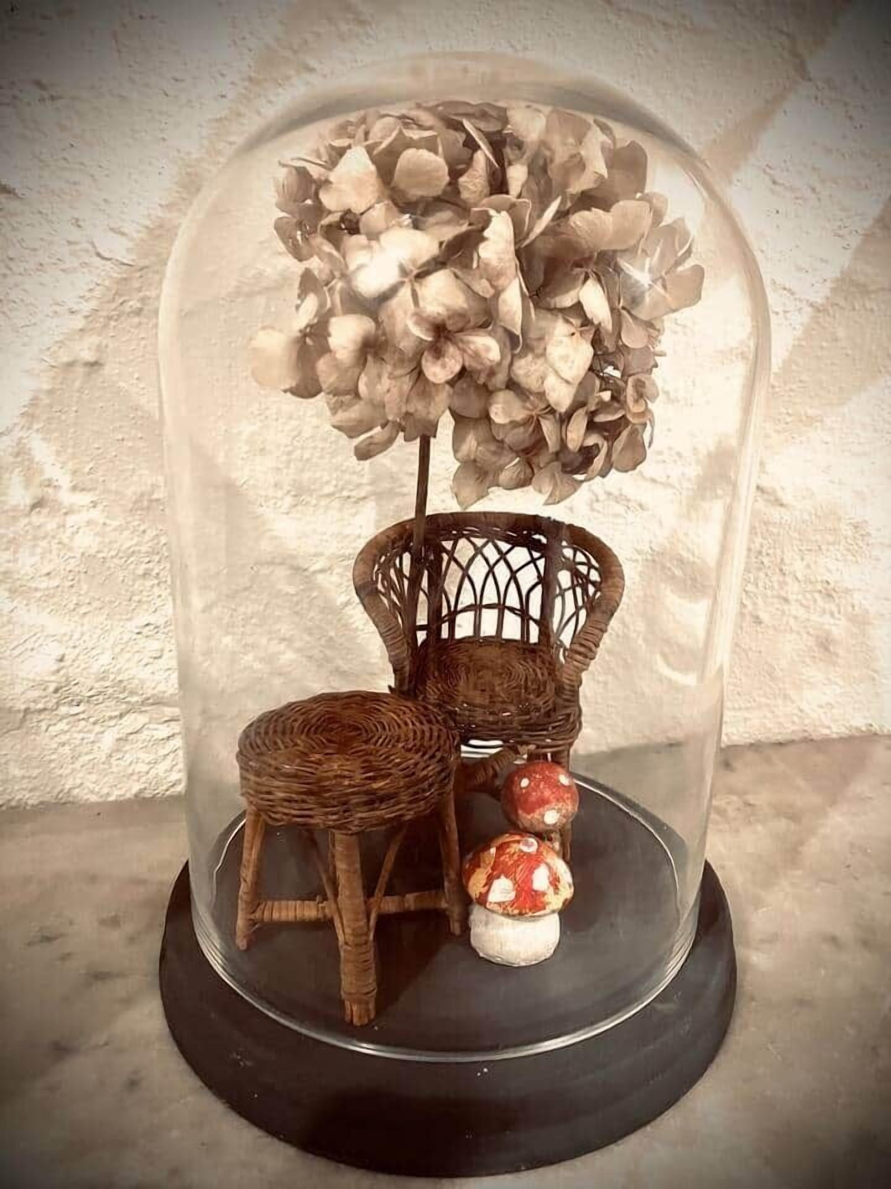 A wicker chair and stool under a glass dome with a floral arrangement.
