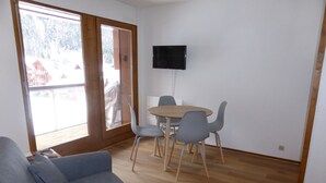 A room with a wooden floor, a grey sofa, a dining table with four chairs, and a flat-screen TV mounted on the wall. - Studio for 4 at Hameaux du Lay with parking (Les Contamines-Montjoie)