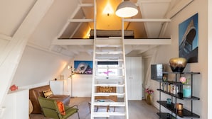 Une chambre à coucher de style loft avec une échelle blanche, une chaise verte, un lit, un bureau, une bibliothèque et un tableau accroché au mur.