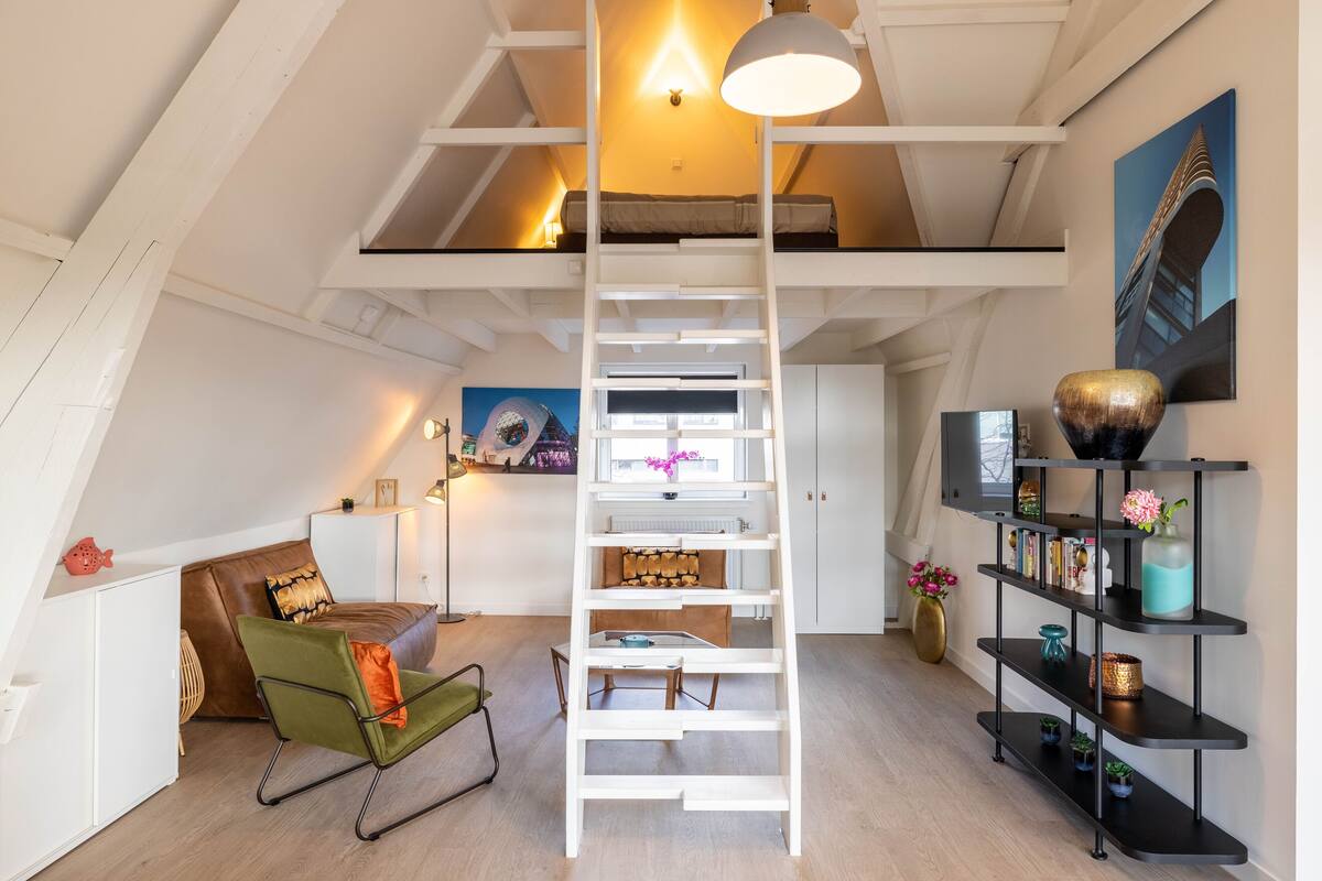 Ein Schlafzimmer im Loft-Stil mit einer weißen Leiter, einem grünen Sessel, einem Bett, einem Schreibtisch, einem Bücherregal und einem Bild an der Wand.