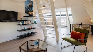 Un salon moderne avec un espace en mezzanine, une bibliothèque, une table basse en verre et un fauteuil vert agrémenté d’un coussin orange.
