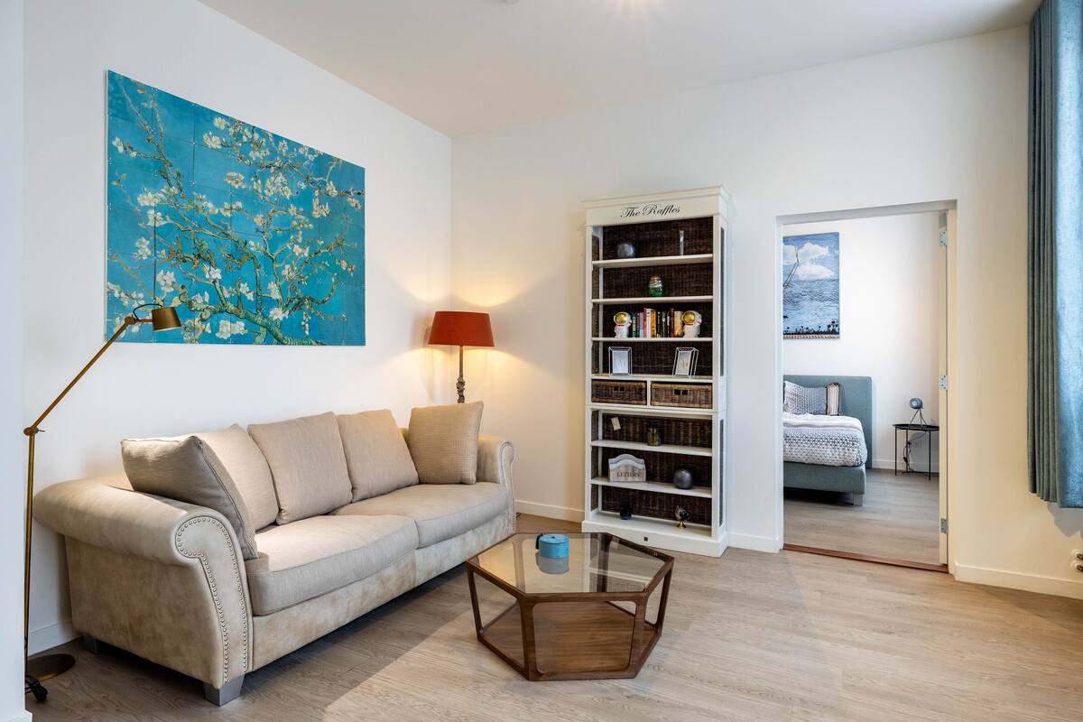 Ein Wohnzimmer mit einem beigefarbenen Sofa, einem Glastisch, einem Bücherregal und einem Gemälde an der Wand.
