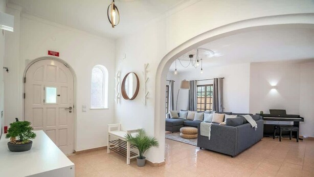 A spacious living room with a grey sofa, a piano, and a white door with a sign. - Spacious House | Terrace & Pool by Host Wise (Lagoa)