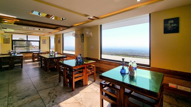 A restaurant with wooden tables and chairs, large windows with a view, and ceiling fans. - Hotel JK Dharamshala (Dharamshala)