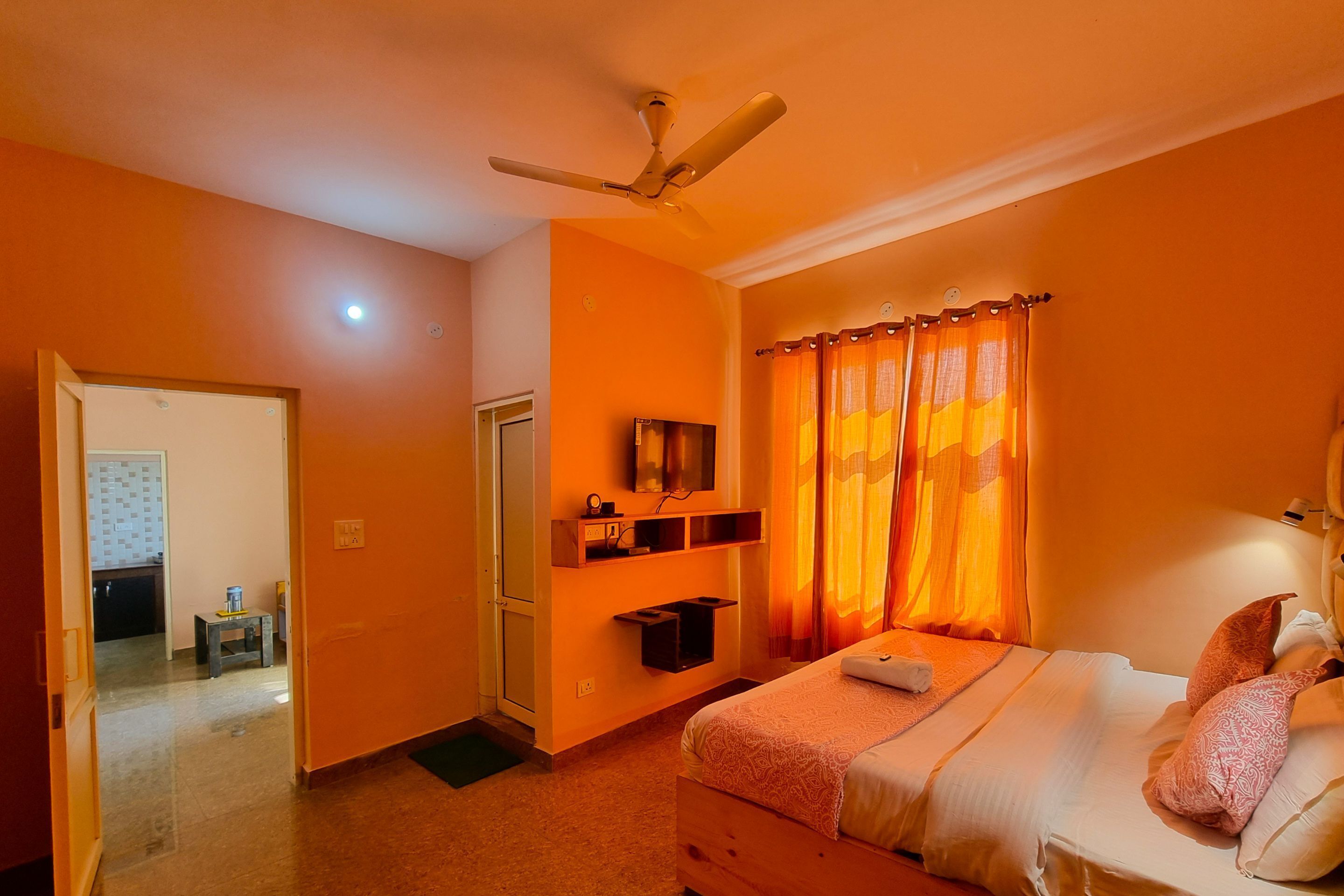 Un dormitorio con cama, ventilador de techo, televisor montado en la pared y ventana con cortinas naranjas.