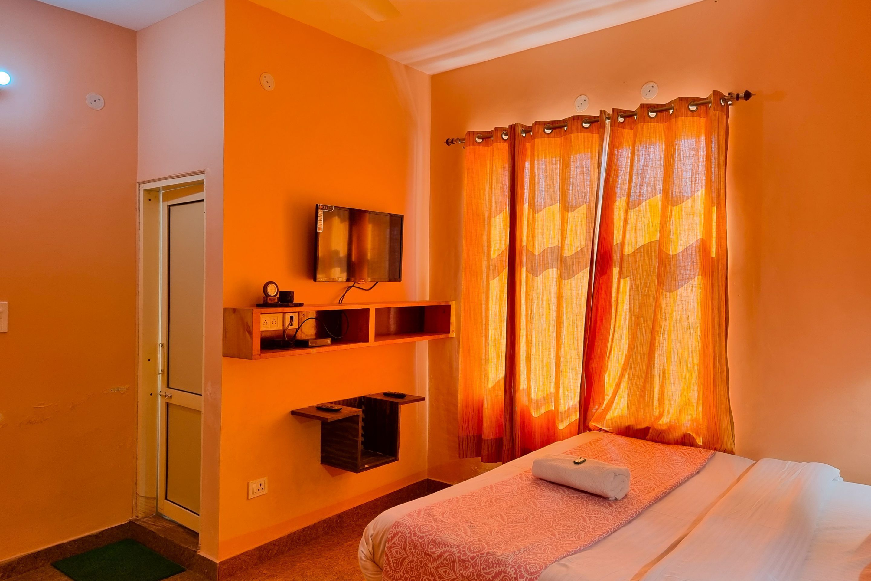 A hotel room with a bed, a TV mounted on the wall, a shelf with a small table, and a door.