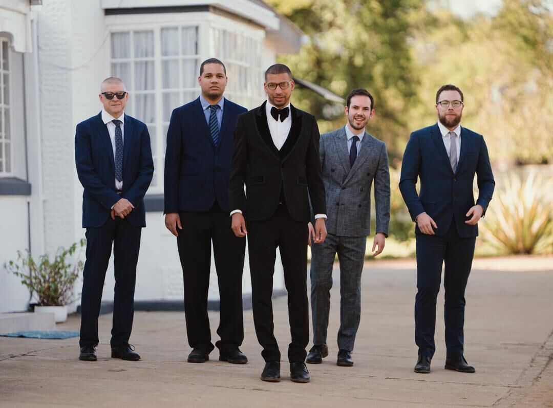 Cinco hombres con traje de pie en fila sobre un camino de hormigón.