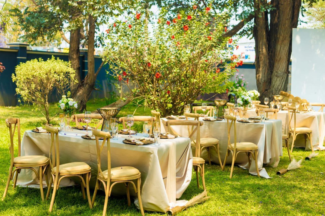 An outdoor dining area with multiple tables set up under trees, each with chairs and table settings.