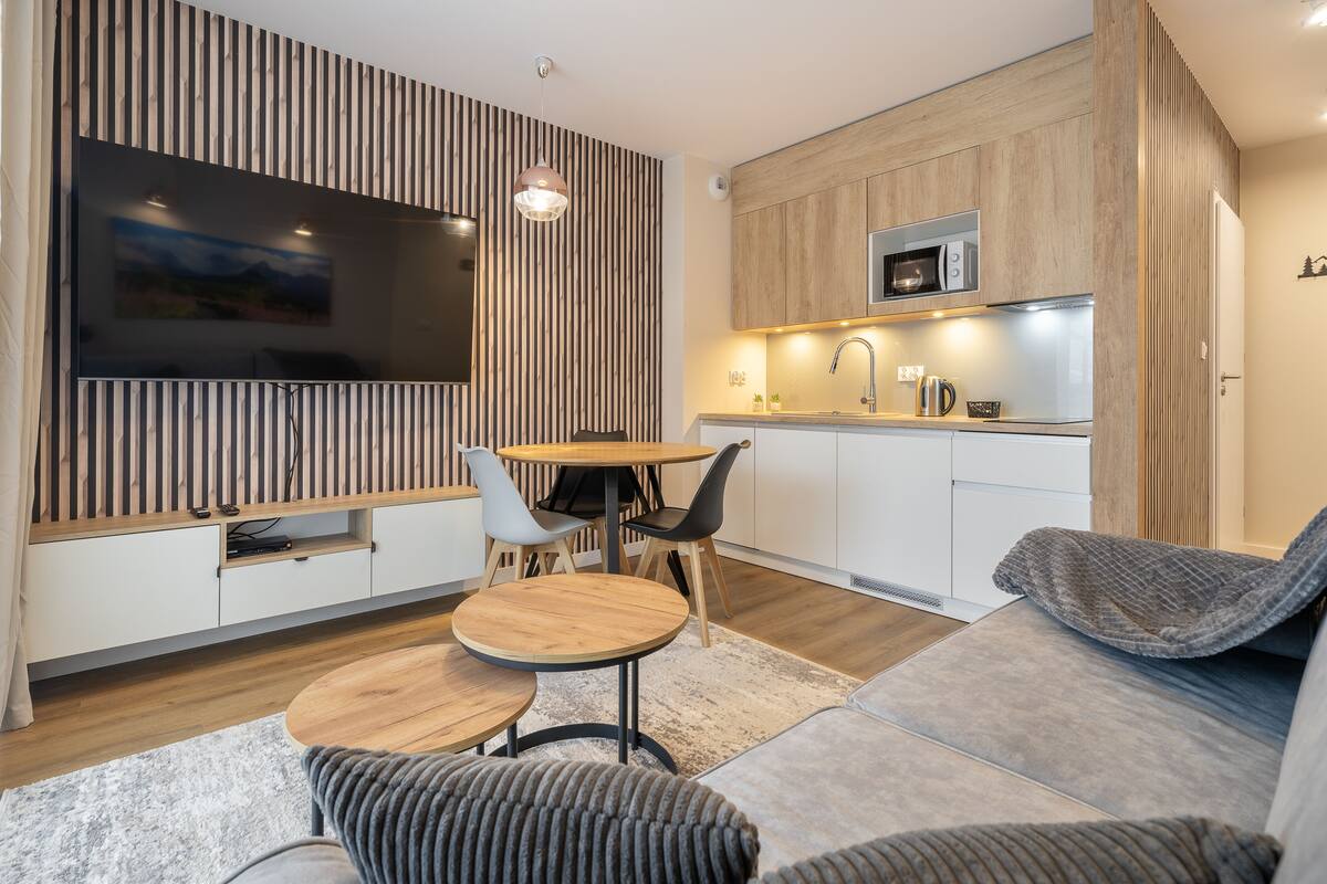 Una sala de estar moderna con televisor de pantalla plana, detalles de madera, un comedor con mesa y sillas redondas, y una kitchenette con microondas.