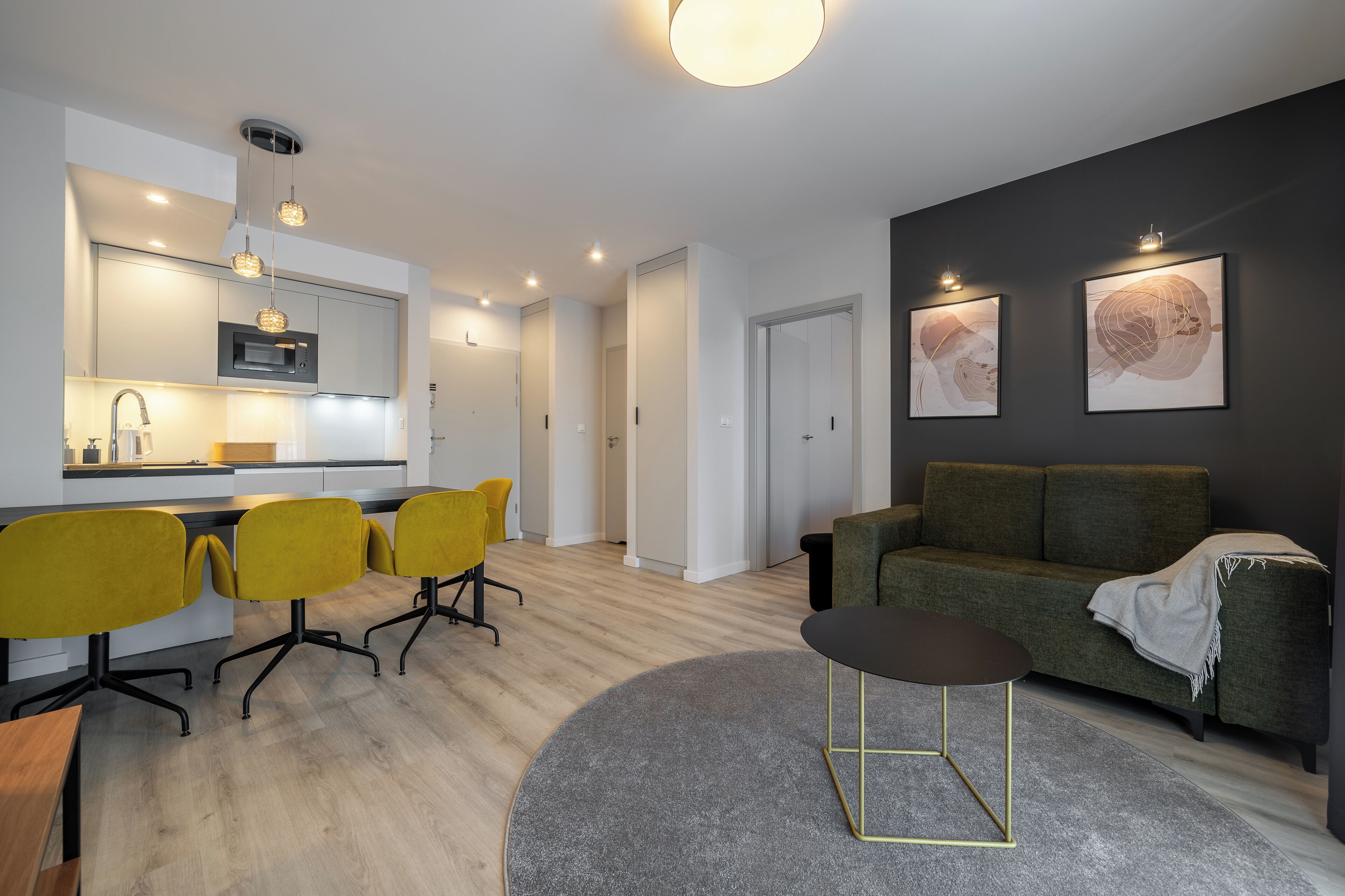 Un salón moderno con un sofá verde, sillas amarillas, una mesa de centro redonda y una zona de cocina con microondas.