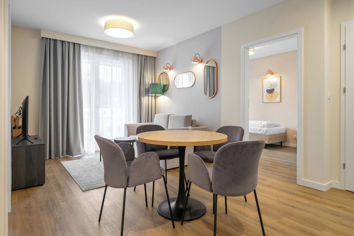 Una sala moderna con una mesa redonda de madera, sillas tapizadas de color gris, un sofá gris y una cama al fondo.