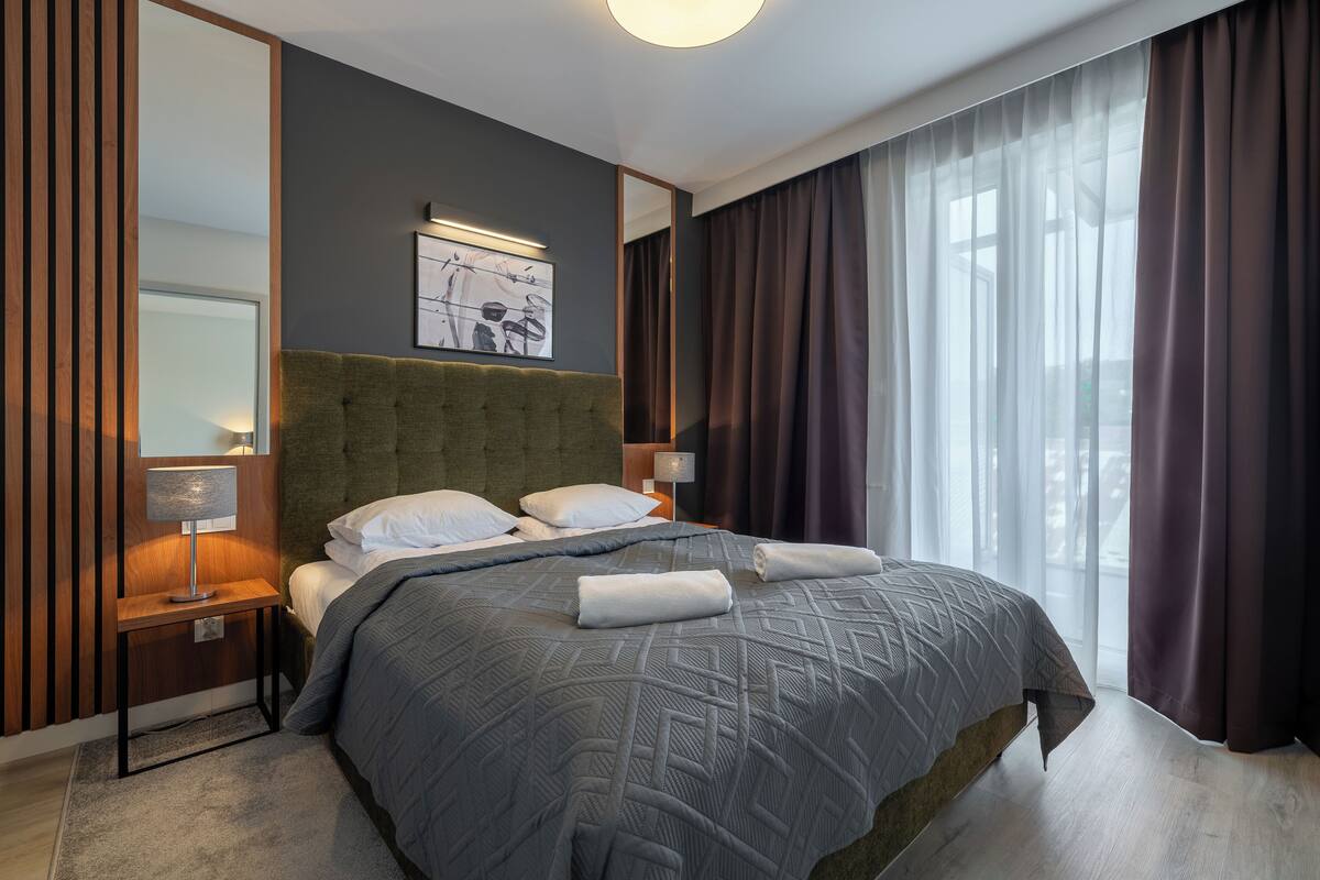 Habitación de hotel moderna con una cama grande, cabecera, mesita de noche con lámpara, una obra de arte en la pared y vistas al exterior a través de cortinas translúcidas.