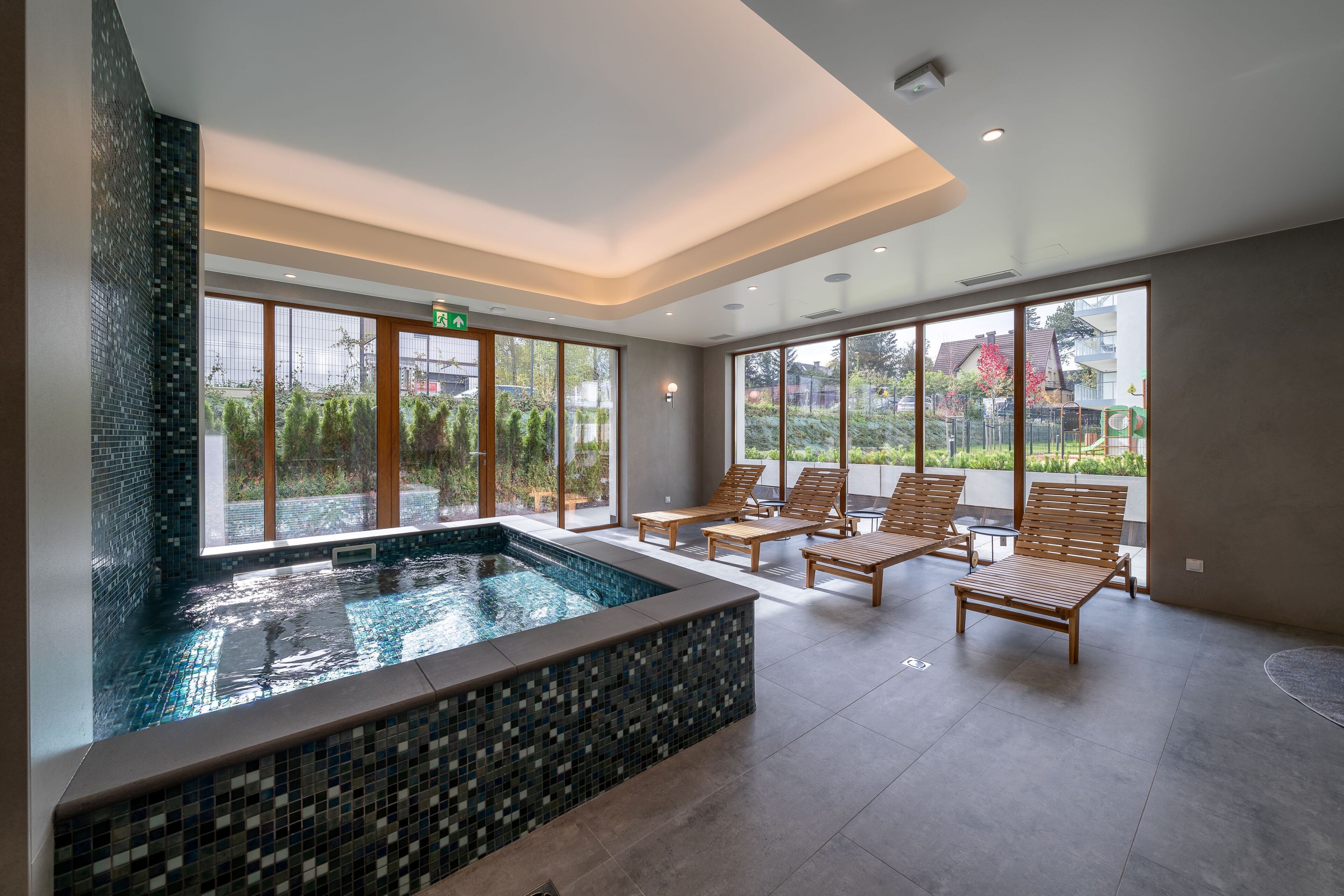 A modern indoor pool area with a tiled edge, wooden lounge chairs, and large windows offering a view of greenery and buildings.