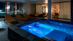 Uma área interna de piscina moderna, com um elemento aquático iluminado em azul, espreguiçadeiras de madeira e uma parede de vidro com vista para o exterior.