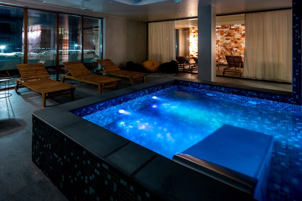 A modern indoor pool area with a blue-lit water feature, wooden lounge chairs, and a glass wall offering a view of the outside. - RentPlanet - Szymony II (Zakopane)