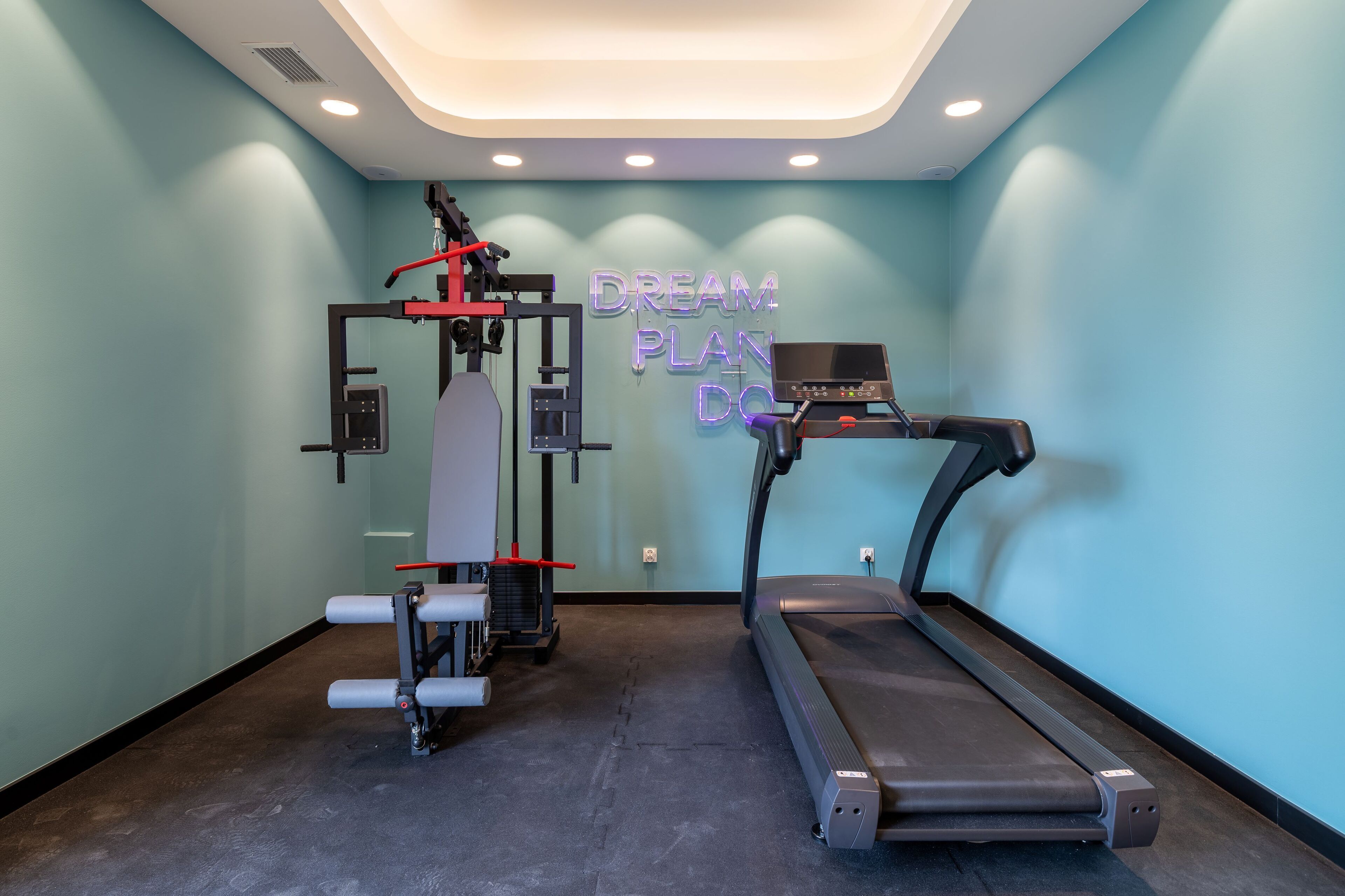 A gym with a treadmill and a weight machine, a neon sign on the wall, and a light blue wall.