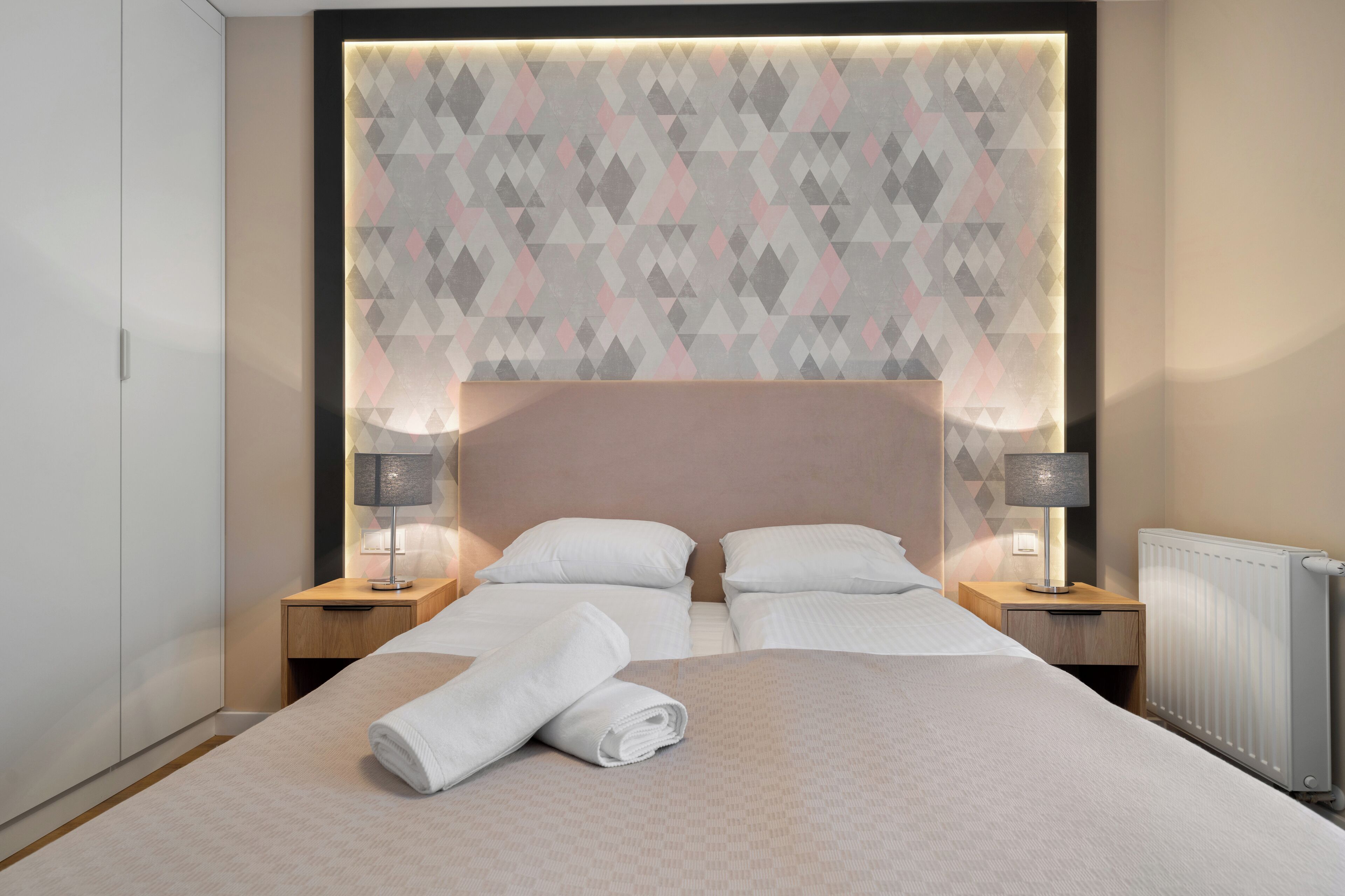 Een keurig opgemaakt bed met witte lakens en een opgerolde handdoek, twee nachtkastjes met lampen, en een hoofdbord met een geometrisch patroon.