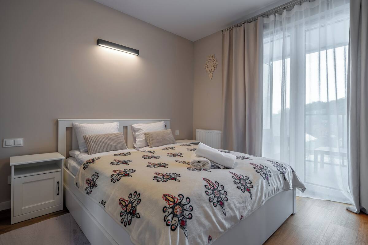 Una cama bien hecha con una colcha de estampado floral, una mesita de noche con una lámpara y una ventana con cortinas traslúcidas.