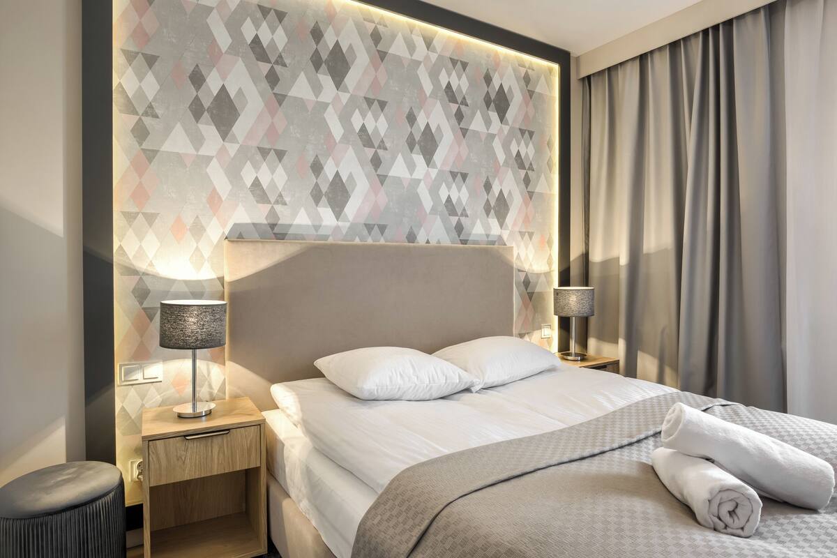 Habitación de hotel con una cama, dos lámparas de noche, una mesita de noche, una pared con estampado geométrico y una ventana con cortinas.