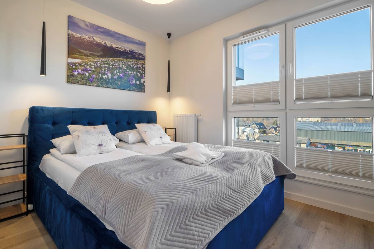 Un dormitorio con una cama grande, vista a la ciudad, un cuadro en la pared y una ventana con persianas.
