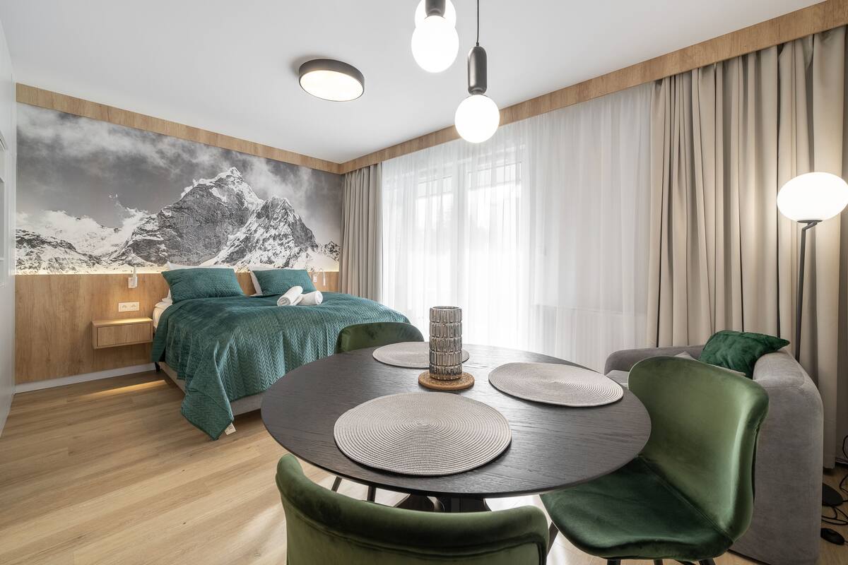 Una habitación de hotel moderna con una cama, una mesa de comedor con sillas, una lámpara y un cuadro grande de un paisaje montañoso en la pared.