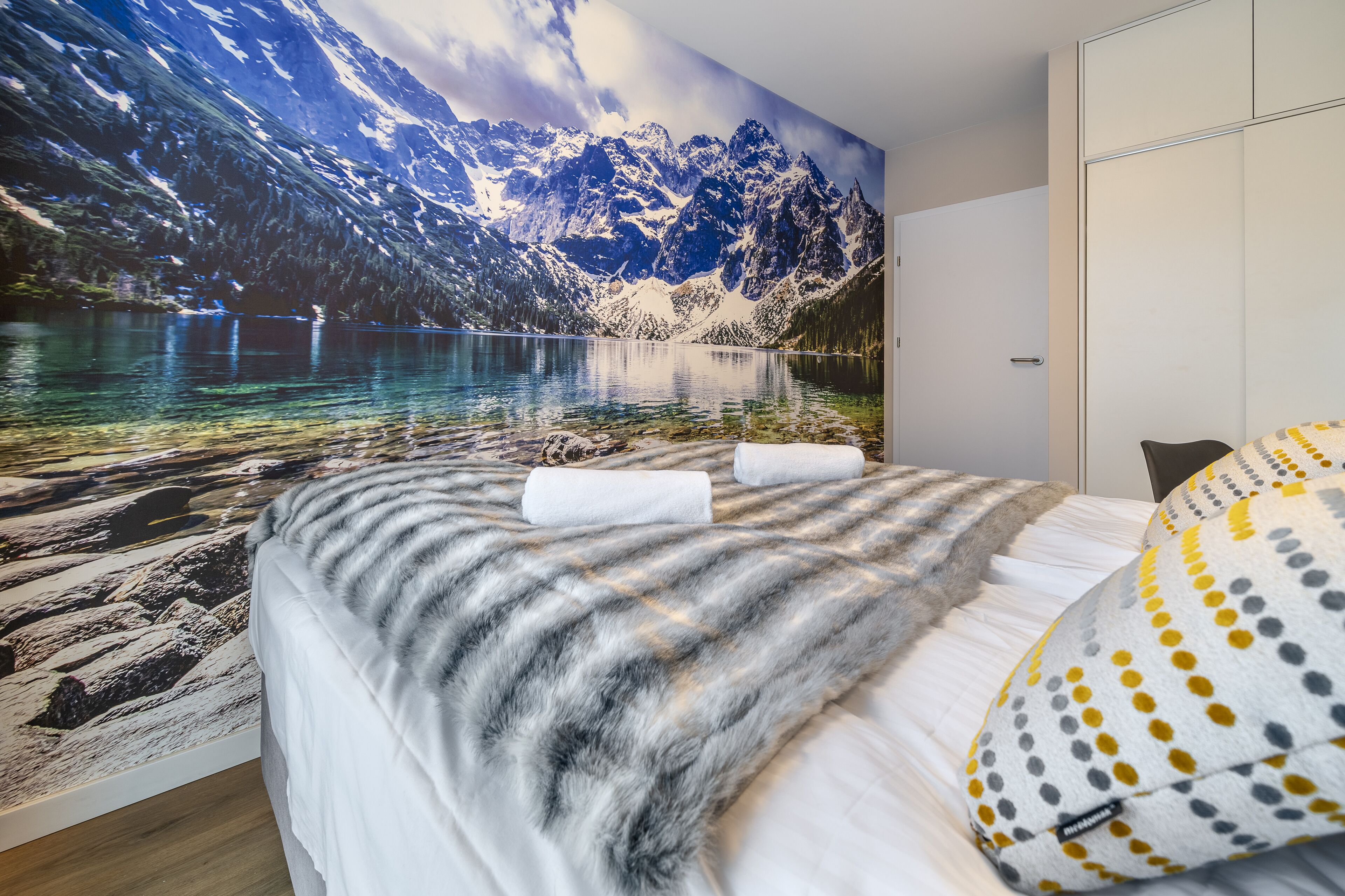 A bedroom with a mountain landscape mural, a bed with a grey and white checkered blanket, and a pillow with yellow and grey polka dots.