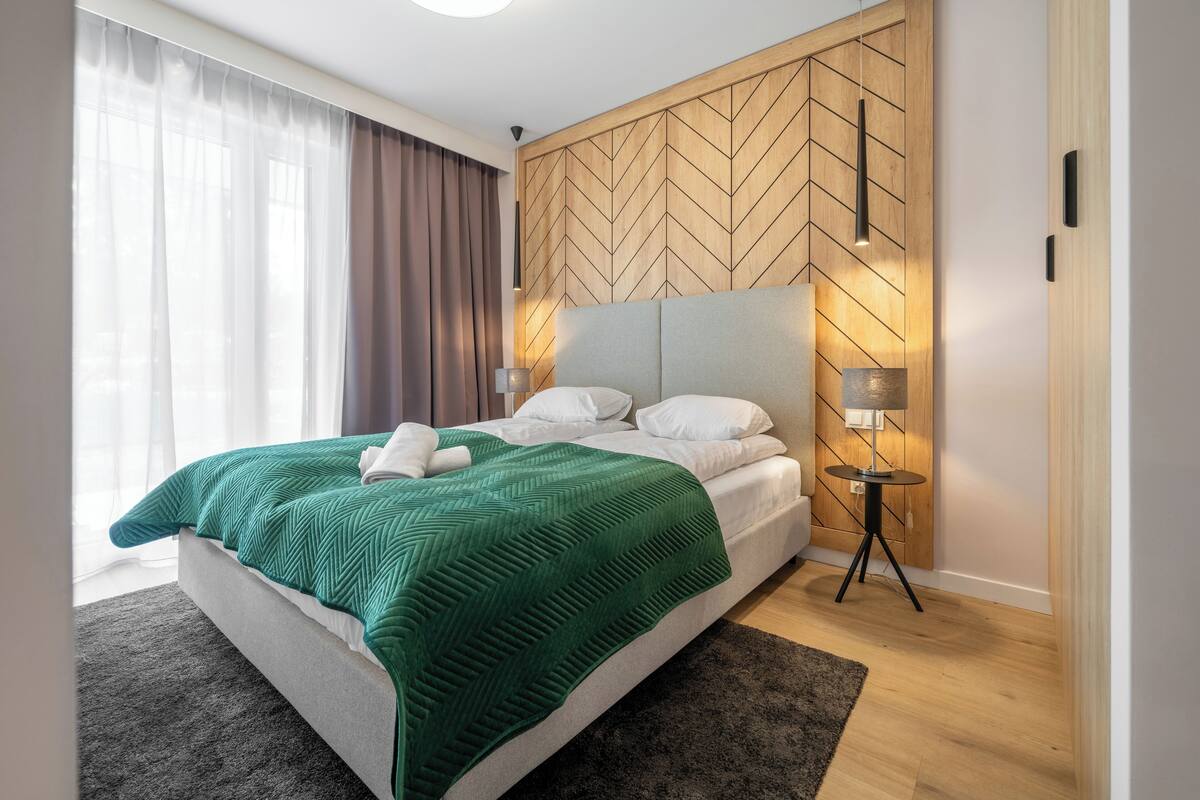 Una habitación de hotel moderna con una cama grande, un cabecero de madera, una mesita de noche con una lámpara y vistas al exterior a través de cortinas translúcidas.