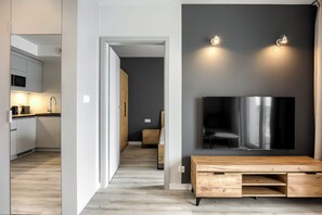 A modern kitchen with a microwave, a wooden cabinet, and a sink. There is a hallway leading to a bedroom with a bed and a dresser. A wooden bench is placed in front of a large flat-screen TV mounted on a dark wall. - RentPlanet - Szymony II (Zakopane)