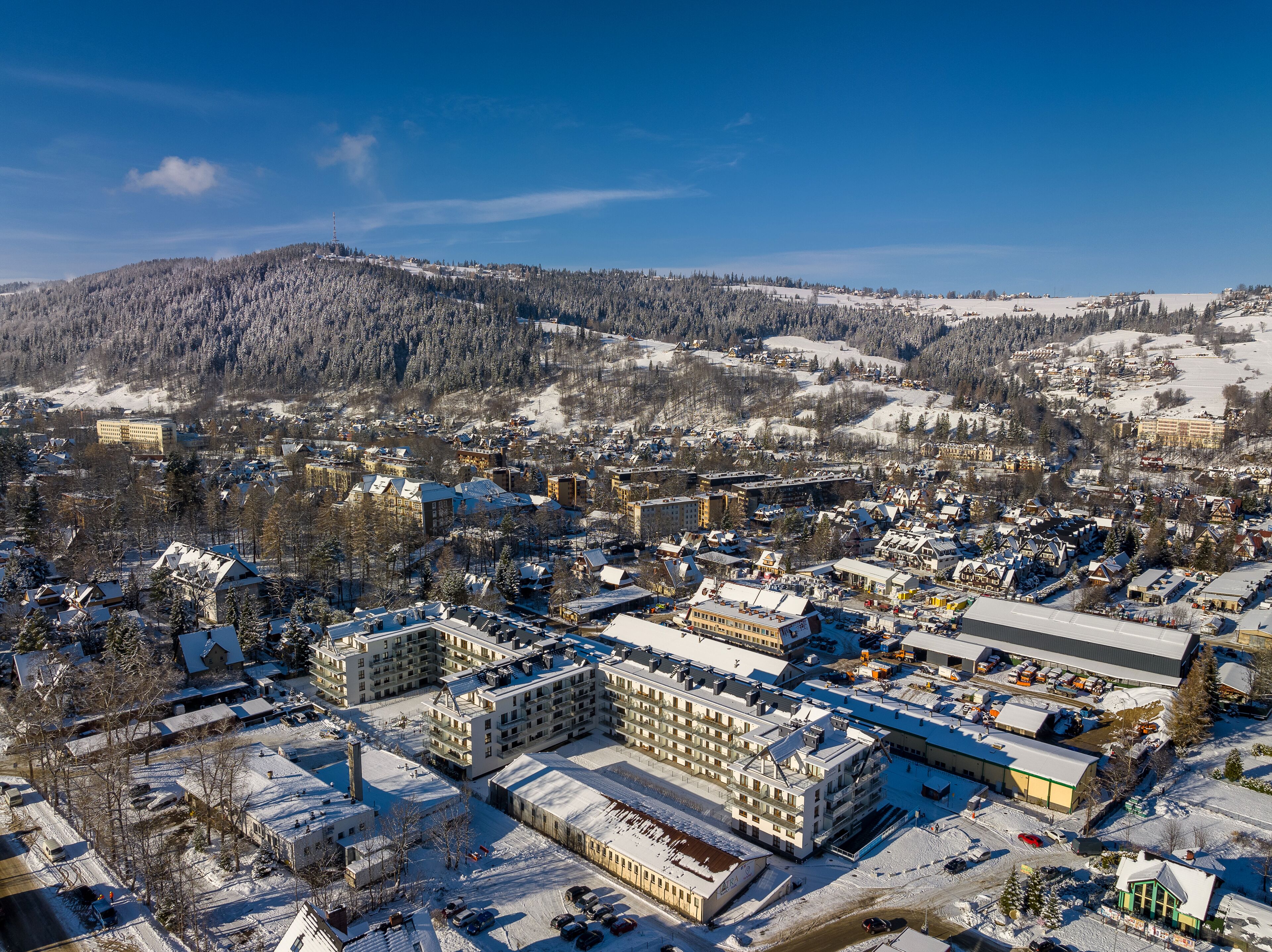 An aerial view of a snow-covered town with buildings, roads, and a forested mountain in the background.