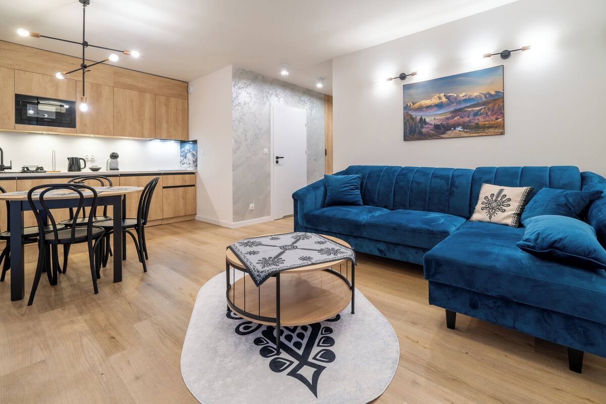 Una sala moderna con un sofá azul, una pequeña mesa redonda y un comedor con sillas, con una cocina al fondo.