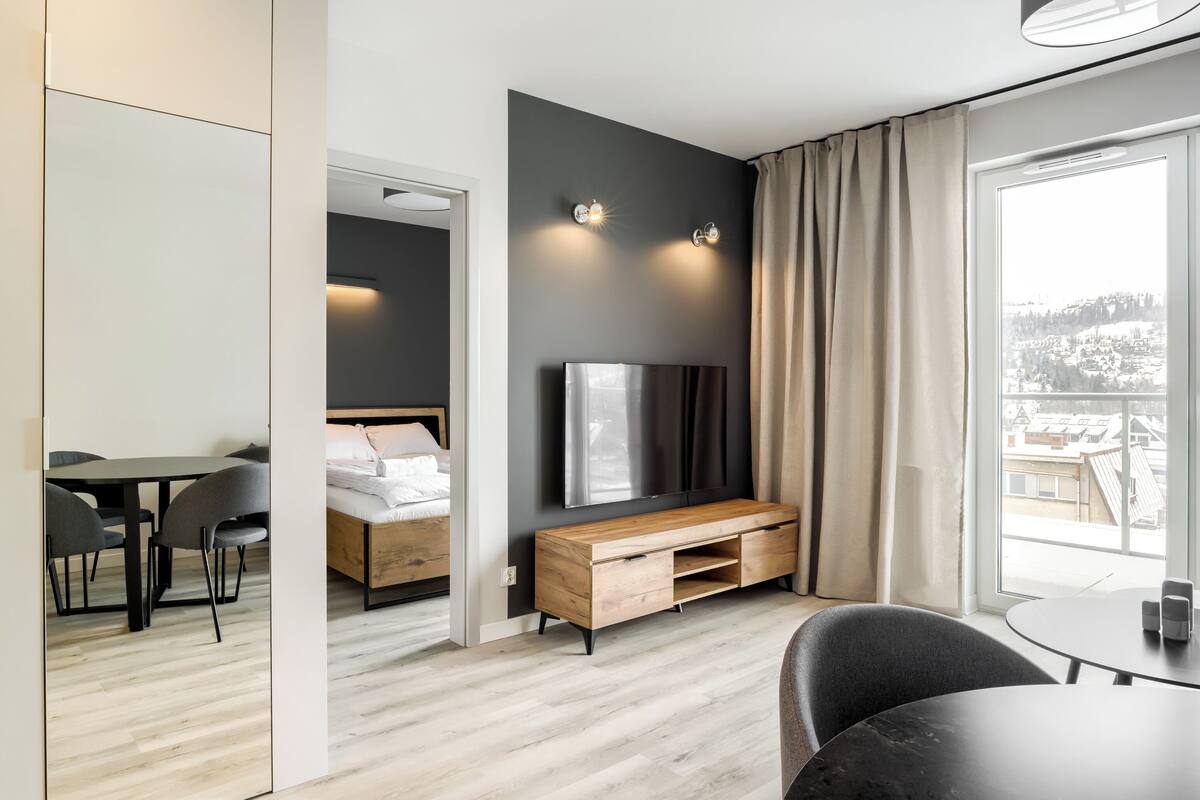 Habitación de hotel moderna con cama, un mueble de televisión de madera, una mesa de comedor y vistas a la ciudad a través de grandes ventanales.