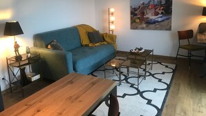 Un soggiorno con un divano blu, un tavolino in legno e un tavolino laterale con piano in vetro. Si vede un quadro appeso al muro e una lampada sul tavolino.