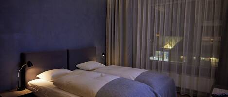 A modern hotel room with a large bed, a bedside table, a lamp, and a view through sheer curtains.