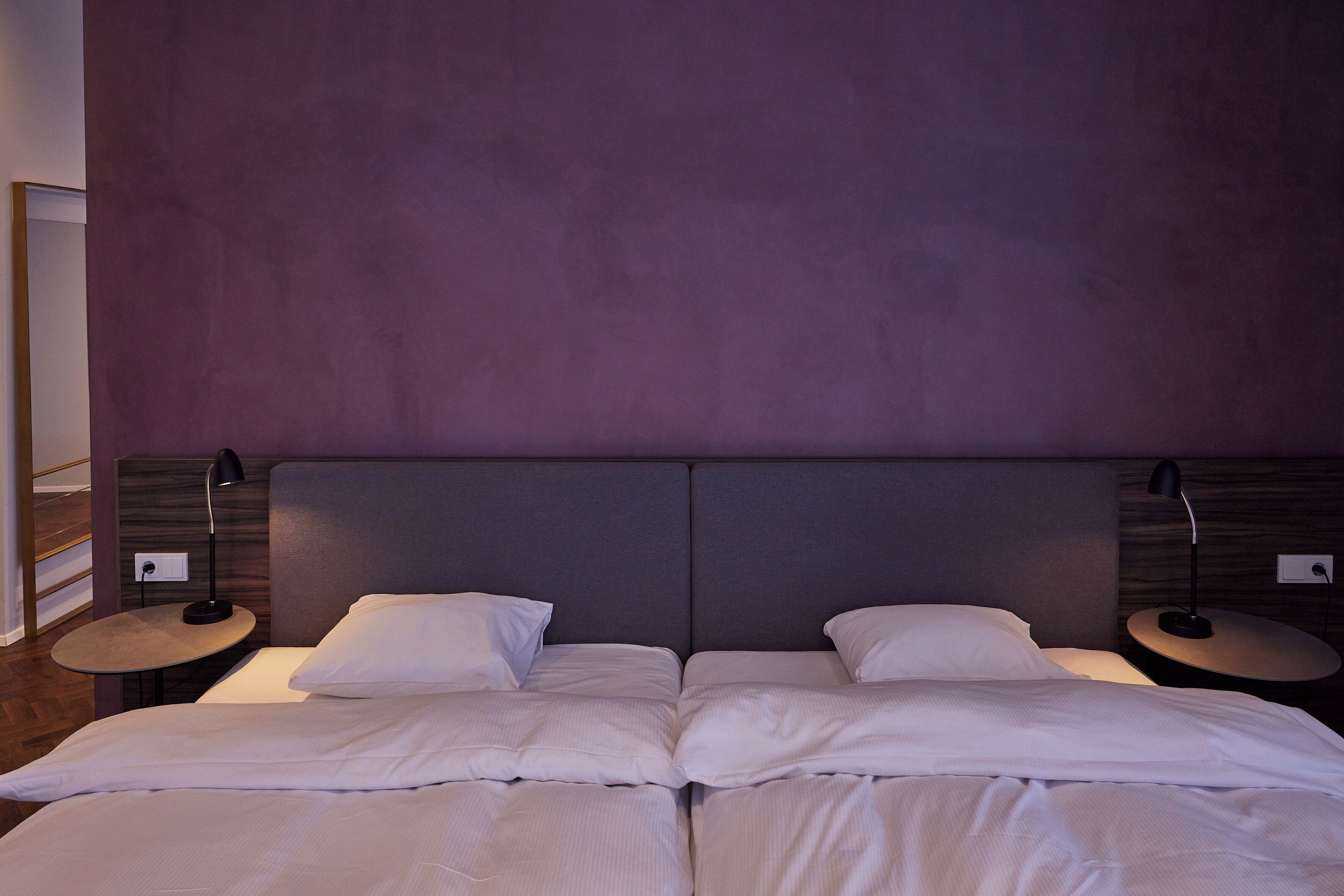 A double bed with white linens, two bedside tables with lamps, and a purple headboard.