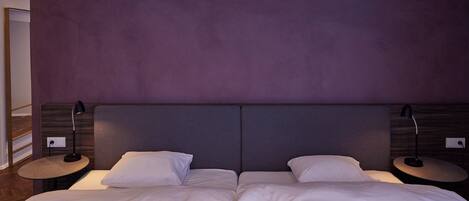 Una cama doble con ropa de cama blanca, dos mesitas de noche con lámparas y un cabecero morado.