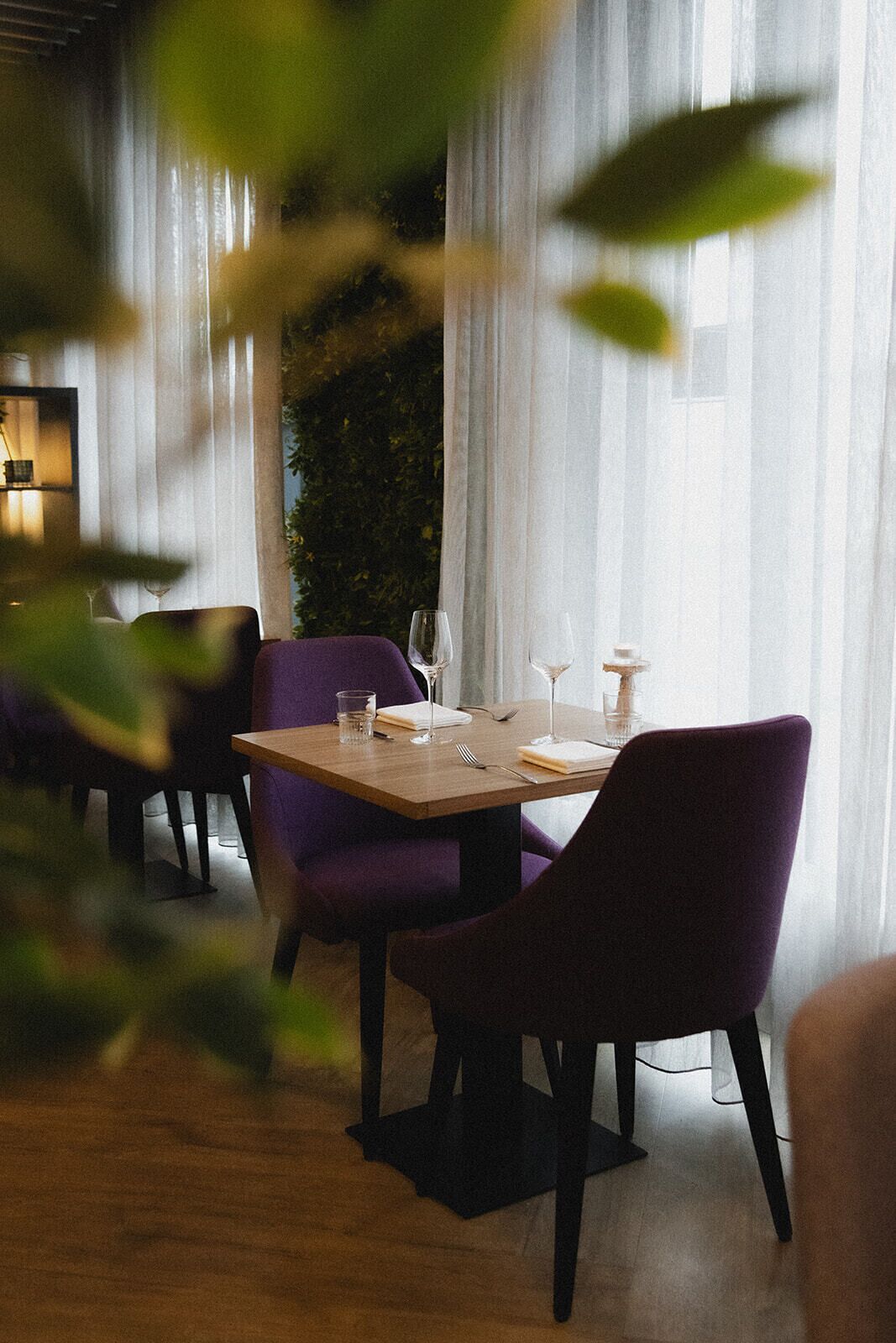 Un coin repas avec une table dressée pour deux personnes, entouré de chaises violettes, et offrant une vue sur la verdure à travers de fines rideaux.