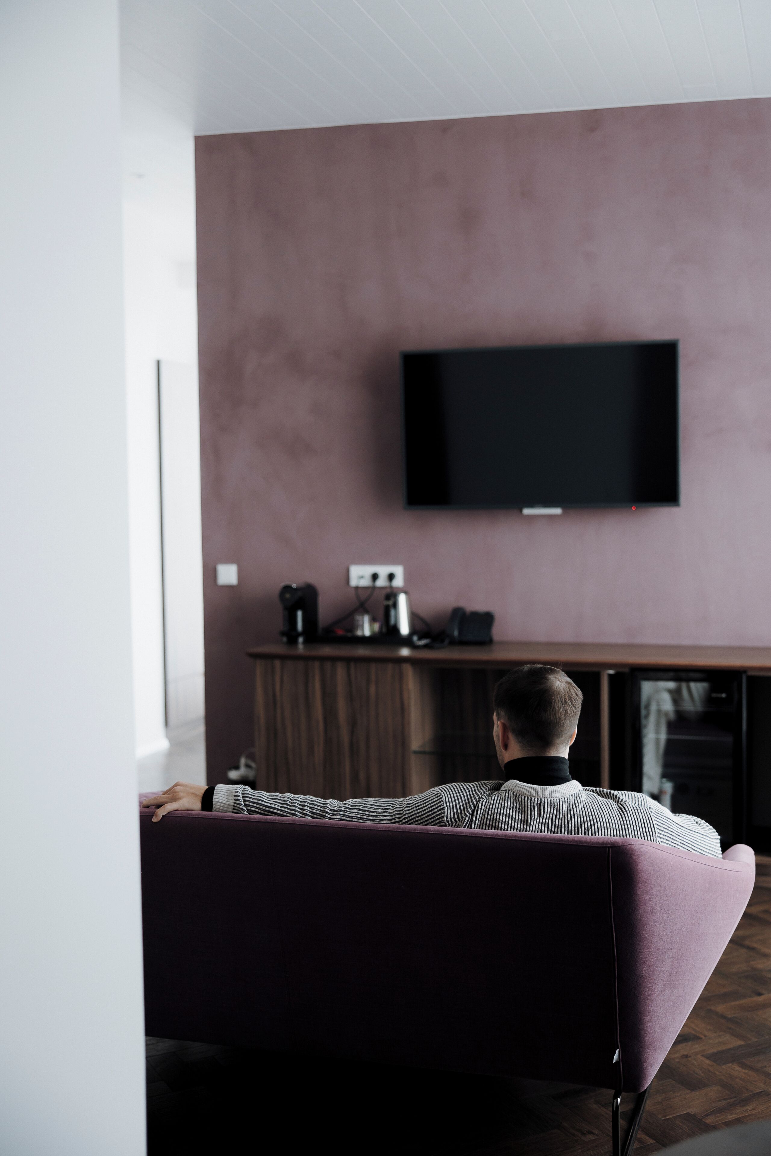 Een moderne woonkamer met een flatscreen televisie aan een wand, een houten kastje eronder, en een persoon die op een bank zit.