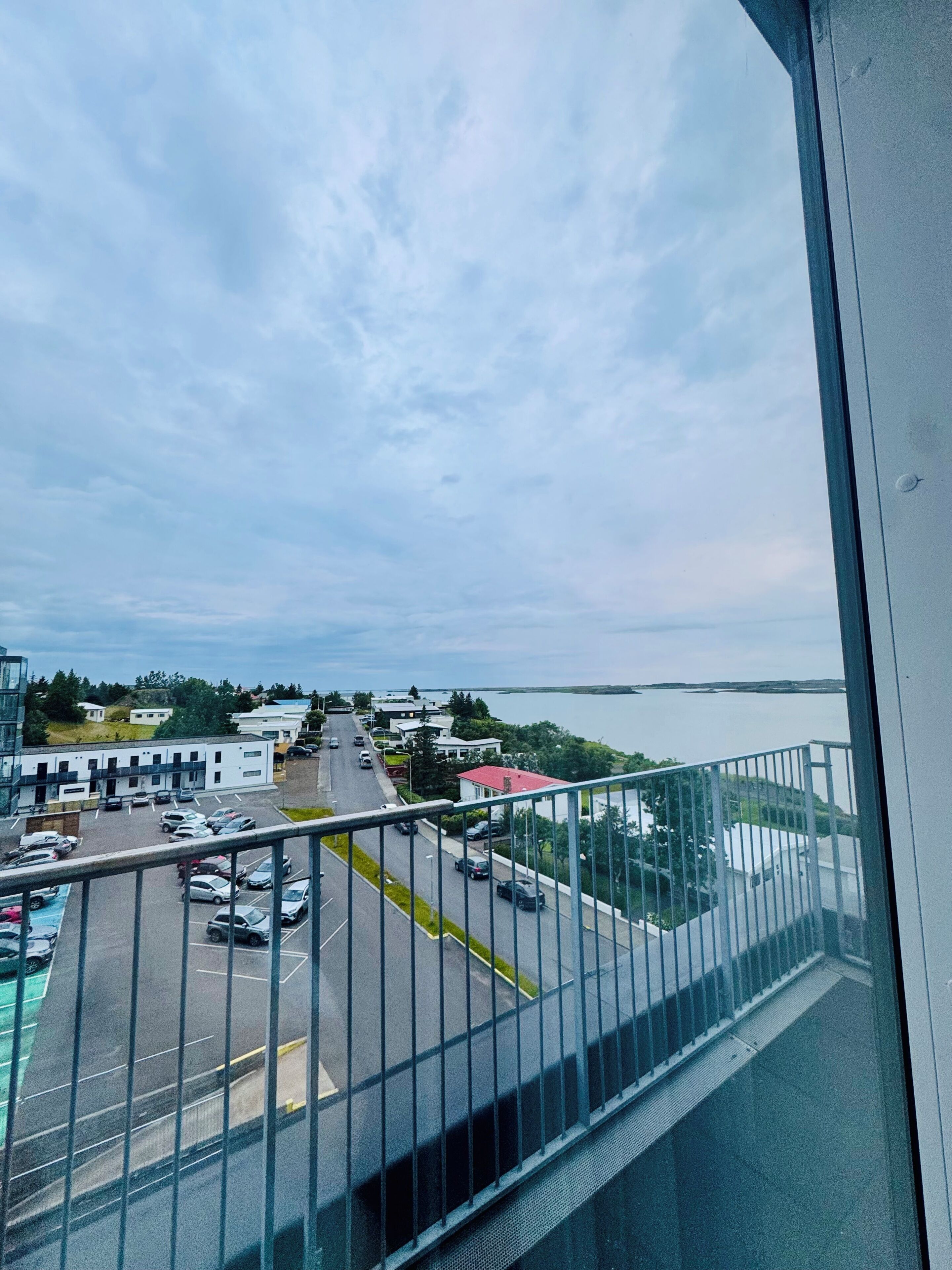 A view from a balcony overlooking a parking lot with several cars, a road, and buildings in the distance.