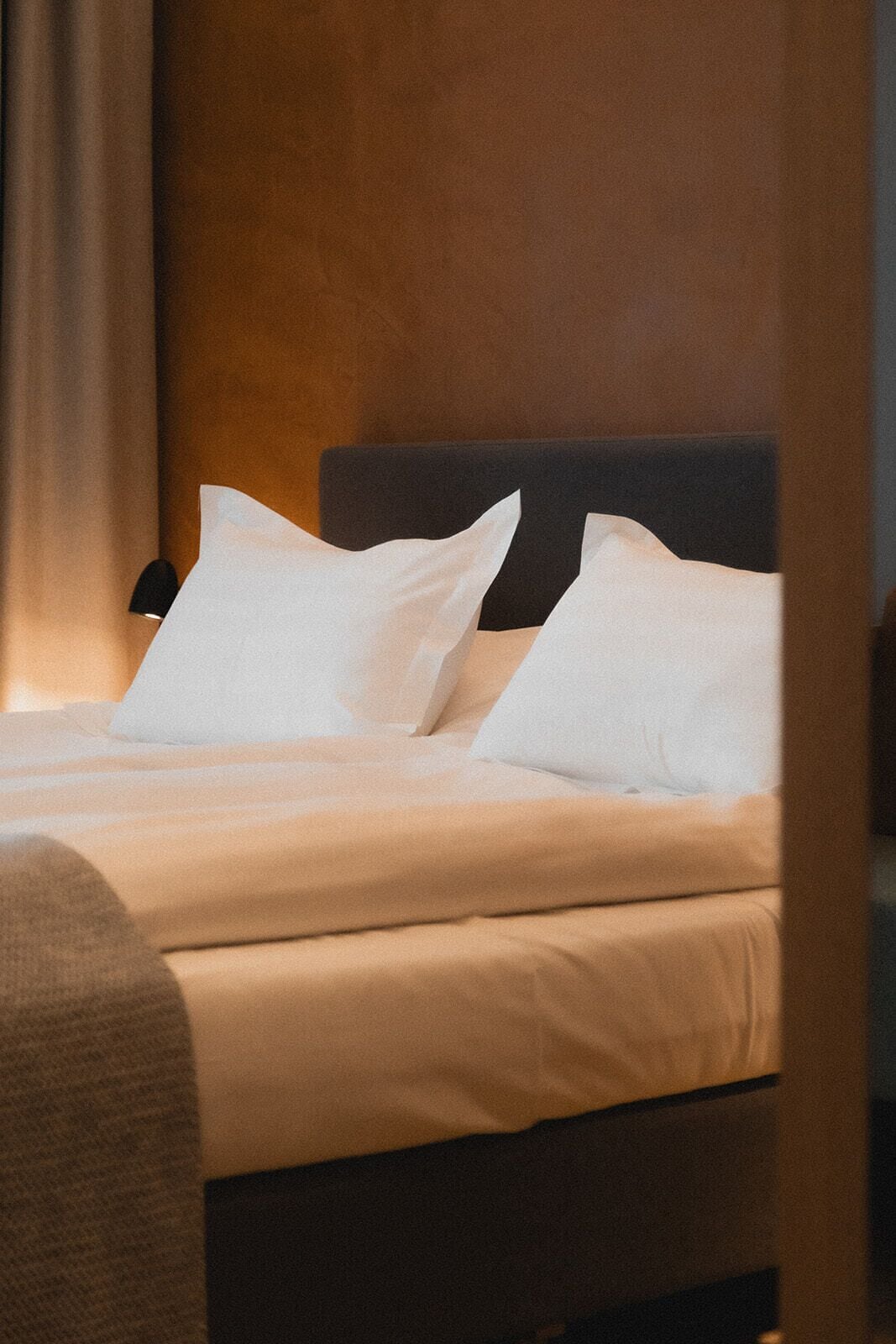 Un lit bien fait, avec du linge de lit blanc et des oreillers, une tête de lit sombre et une partie visible d’un mur en bois.