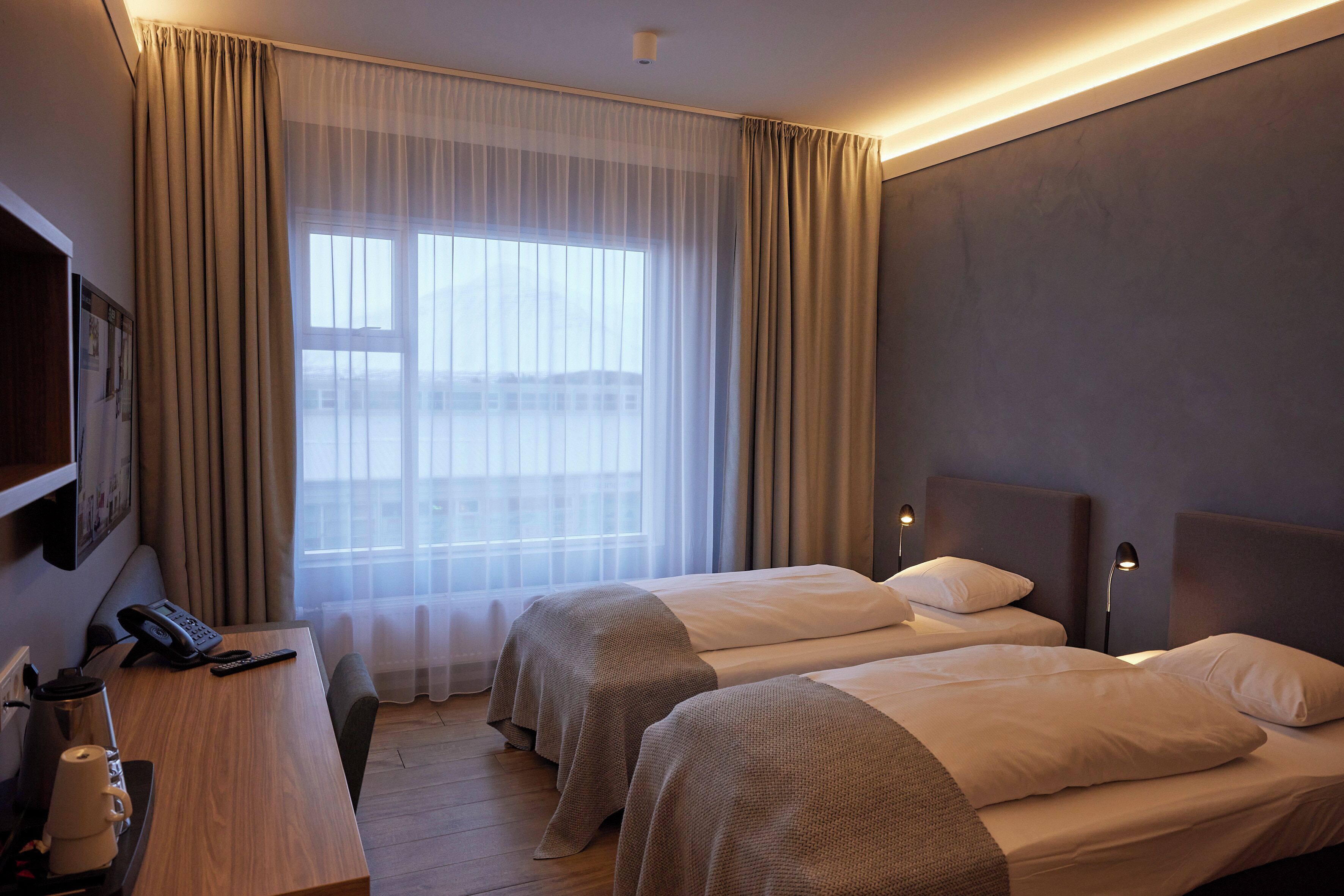 A hotel room with two beds, a desk with a phone, a lamp, and a window with curtains.