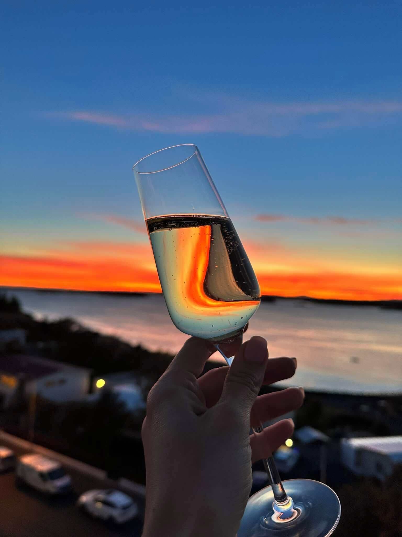 Een hand die een glas wijn vasthoudt, met een zonsondergang op de achtergrond.