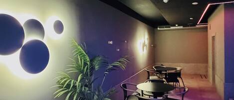 A modern interior with a dining area featuring round tables and chairs, a potted plant, and wall-mounted lighting.