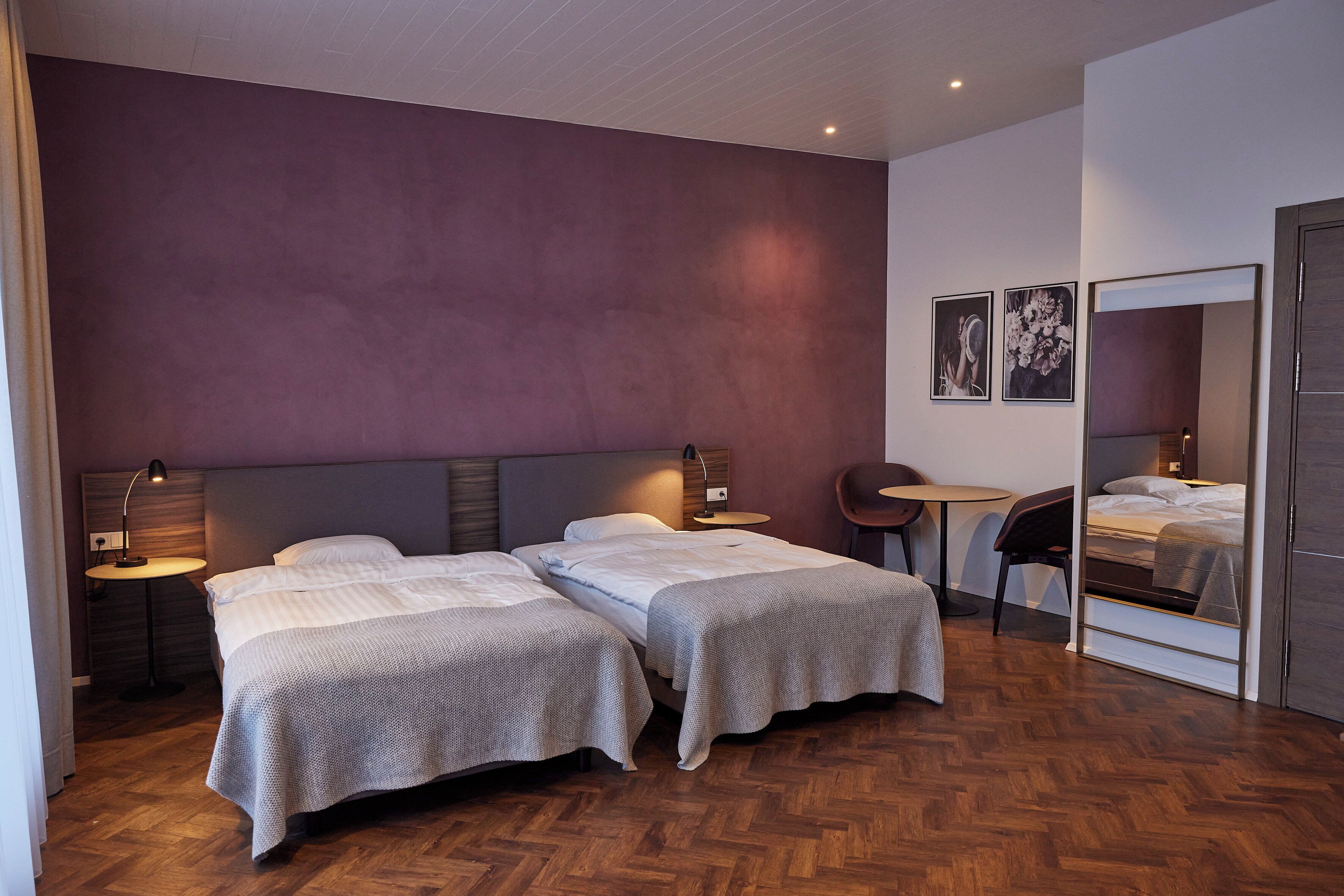 Une chambre d’hôtel avec deux lits, une petite table ronde, une chaise, un miroir et un mur orné de deux tableaux encadrés.
