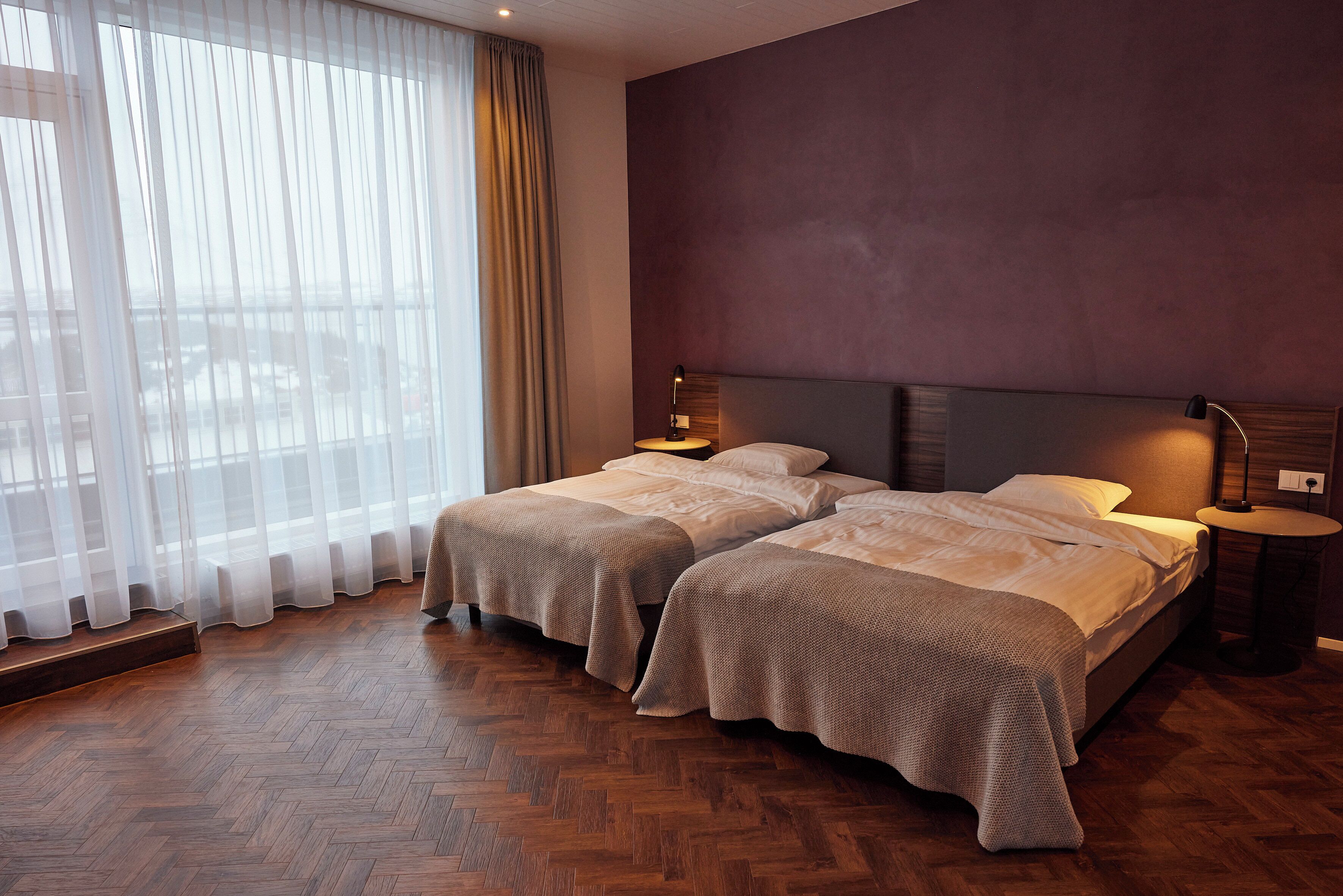 A hotel room with two beds, a large window with sheer curtains, a wooden herringbone floor, and a modern headboard.