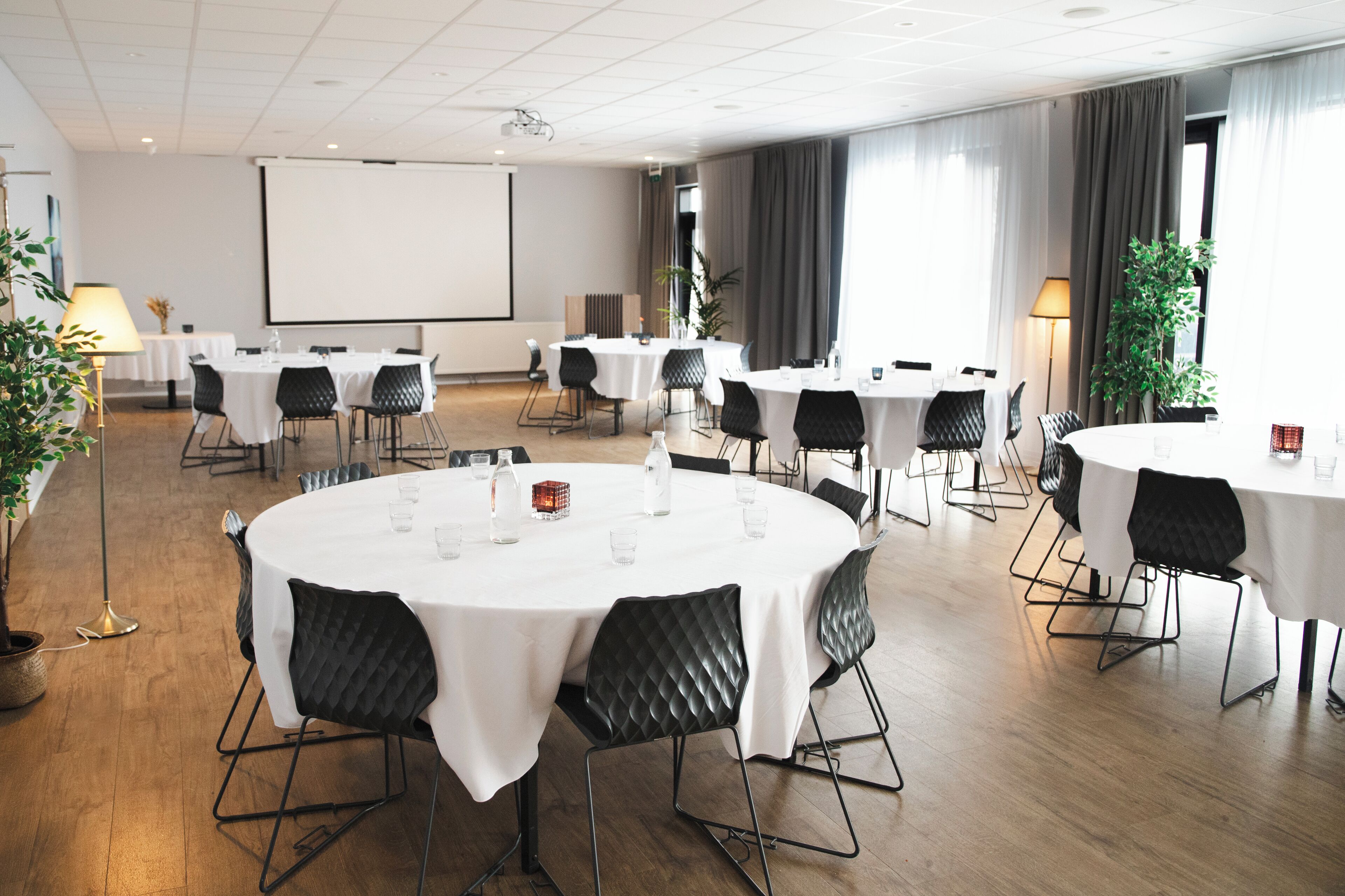 Une salle de conférence équipée de plusieurs tables rondes, de chaises noires et d’un écran de projection.
