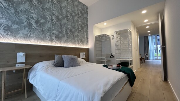 A modern hotel room with a large bed, a bedside table, a mirror, a walk-in closet, and a view of the outside.