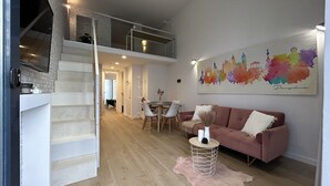 A spacious living area with a high ceiling, a staircase leading to an upper level, a dining area, and a colorful cityscape painting on the wall.