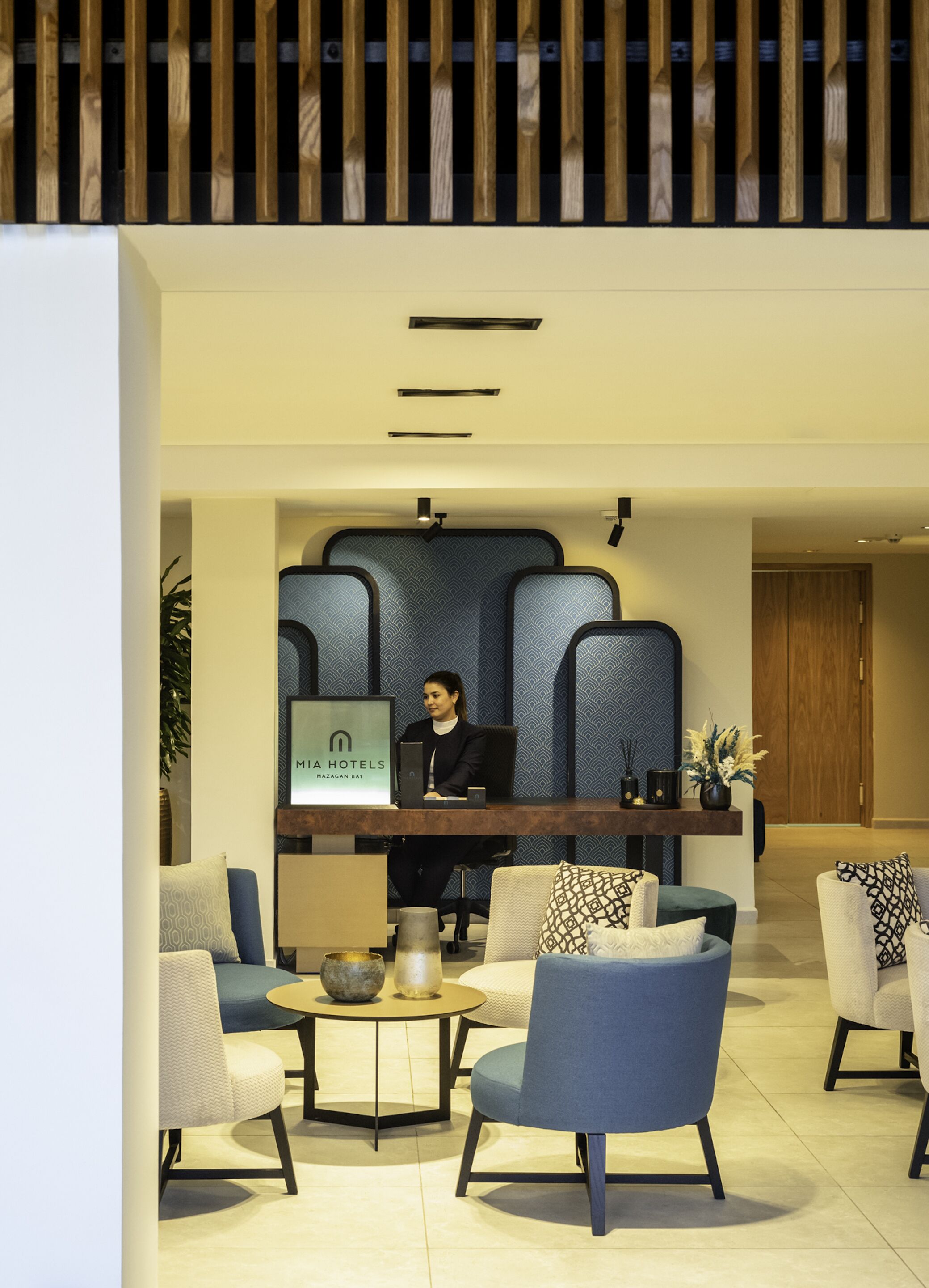 Eine moderne Hotelhalle mit Empfangstresen, Sitzgelegenheiten und einem großen Bildschirm, der den Namen des Hotels anzeigt.