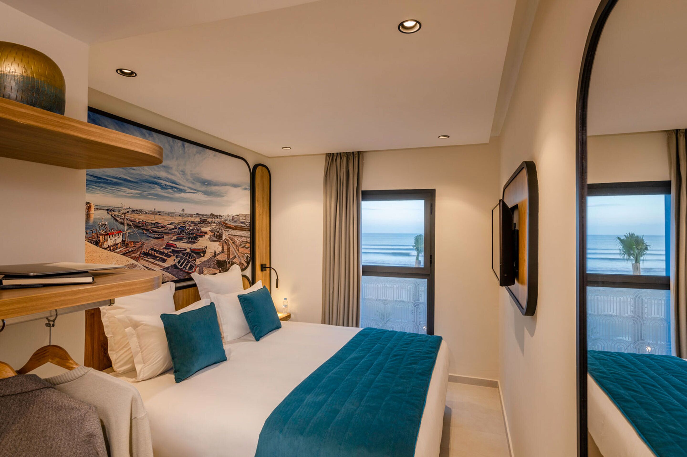 Ett hotellrum med en stor säng, ett skrivbord med en bärbar dator, en tavla som föreställer en kuststad, ett fönster med utsikt över havet och en balkong med en växt.