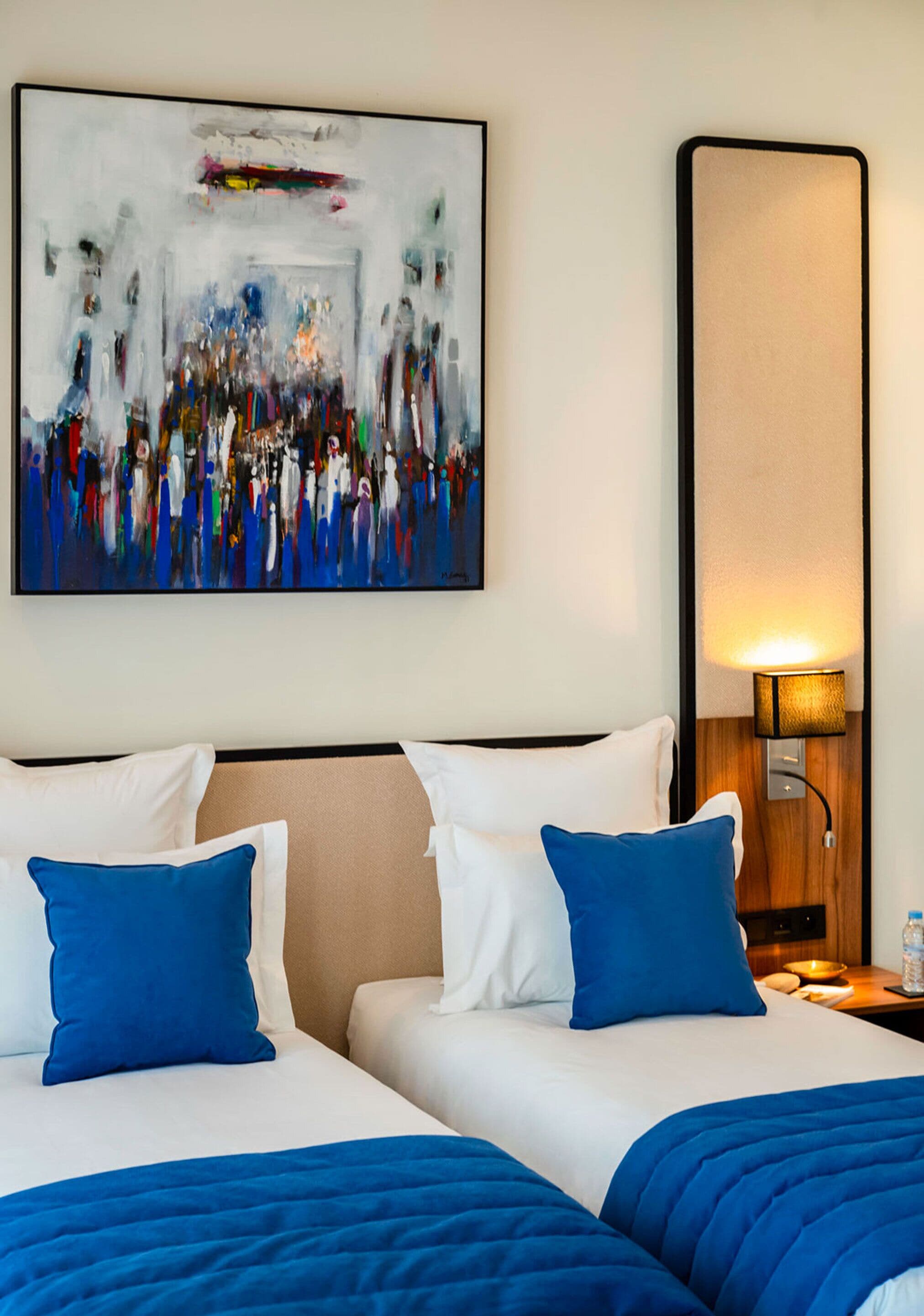 Ein Hotelzimmer mit zwei Betten, jeweils mit weißer und blauer Bettwäsche, einem großen abstrakten Gemälde an der Wand und einer Nachttischlampe.
